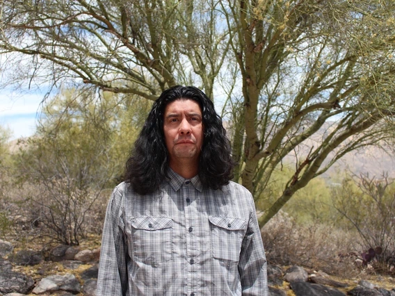 A man with long, wavy black hair stands outdoors in a desert-like environment with green trees and dry, rocky terrain in the background. He wears a buttoned-up, plaid long-sleeve shirt and has a serious expression on his face. The sky is bright and clear, with soft natural lighting casting shadows on the landscape