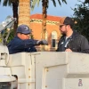 Two University of Arizona Facilities Management workers talk beside a bucket truck on campus. One worker sits inside the raised bucket, wearing gloves and a U of A–branded cap, while the other stands beside the truck, listening. Old Main is visible in the background along with palm trees.